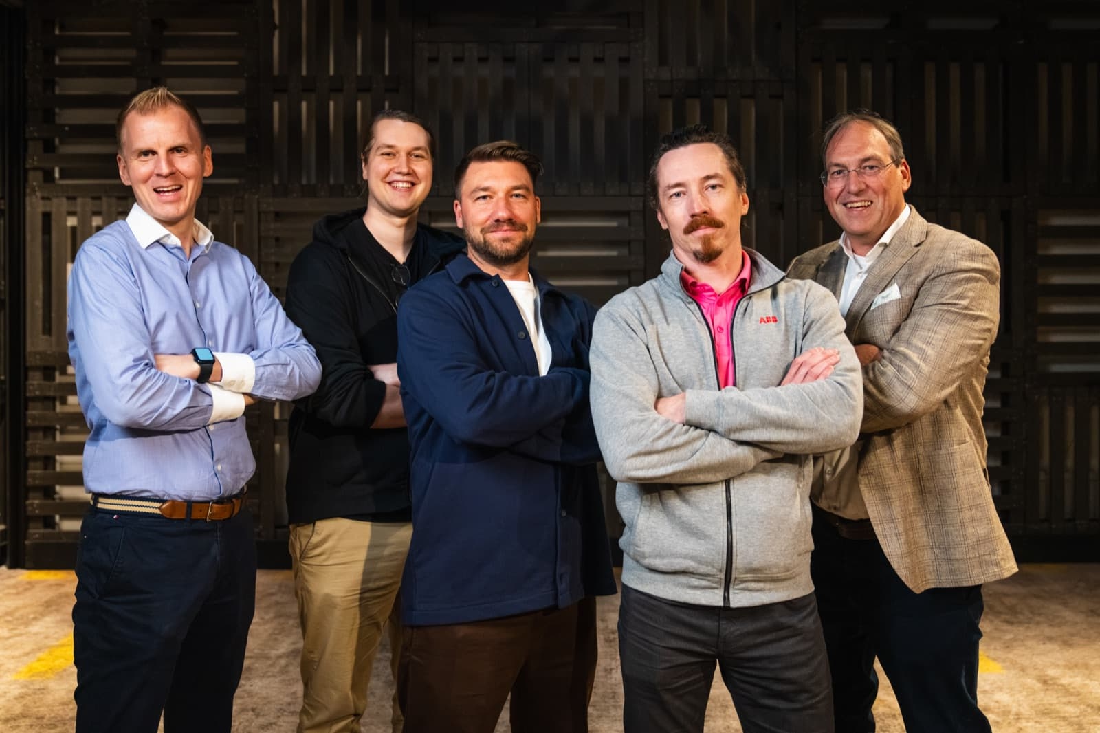 Five men standing side by side, most with their arms crossed, in front of a dark slatted wooden wall.